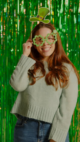 Vertikales-Video-Einer-Frau,-Die-Den-St.-Patrick&#39;s-Day-Feiert-Und-Vor-Einem-Grünen-Lametta-Vorhang-Steht-Und-Eine-Brille-In-Form-Eines-Kleeblatts-Trägt